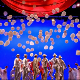 large crowd of performers standing on a stage while white ballons fall to the ground